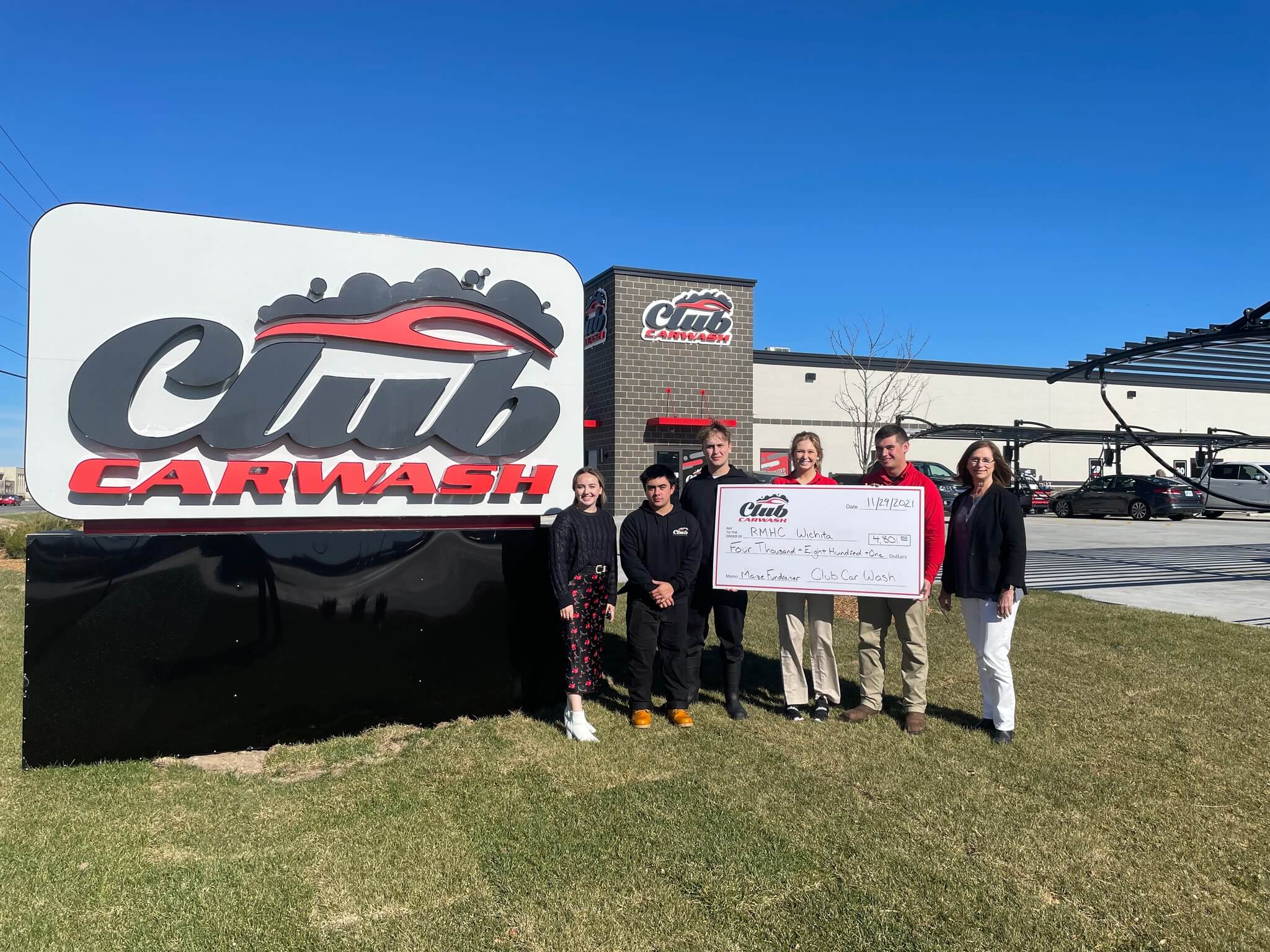 Group of Club Car Wash Employees and representatives from the Ronald McDonald House holding a check in the grass next to Club Car Wash entry sign.